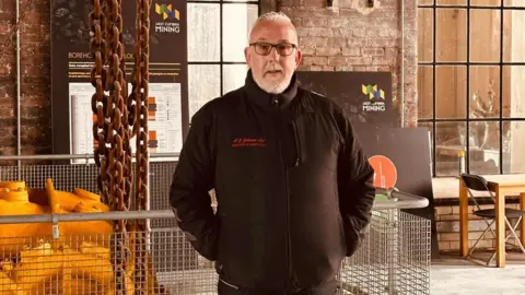 Mike Johnson Mike Johnson, owner of the Haig Colliery Mining Museum, faces the camera. Behind him there are large metal chains, part of mining machinery that is in the engine hall at the Haig Colliery in Whitehaven, West Cumbria.