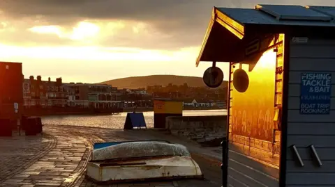 Robin Red Breast A sunset scene of a harbour with a hut on the right hand side in the foreground, and a crescent of buildings around a seafront behind. The hut is lit up by the setting sun.