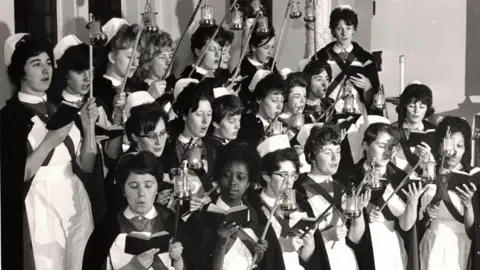 Alistair Ritch A black and white image of 21 female nurses singing carols in the 1960s. They are stood in three descending rows with song books open. The nurses are in uniforms with white collars and pinafores, and wearing white hats. They all carry lanterns.
