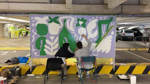 Two people sat down on chairs painting a canvas inside a car park - it has green white and purple coloured designs on it.