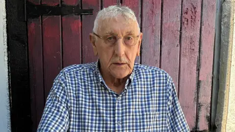 Local Democracy Reporting Service Eddie Wood is stood in front of a red wooden door. He is wearing a chequered blue shirt. He wears glasses and has a moustache and short grey hair. He is looking straight at the camera with a straight facial expression.