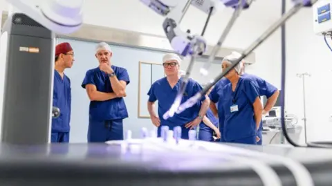 The Royal Wolverhampton NHS Trust Equipment is in the foreground and the four people are in health uniform in the background, along with two others whose faces are not visible.