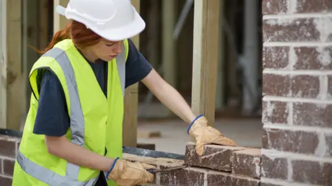 A woman doing bricklaying. She wears a white hard hat and a green high-vis jacket with brown gloves and holds a metal instrument with the tip just under a brick which she is lifting with her left hand.