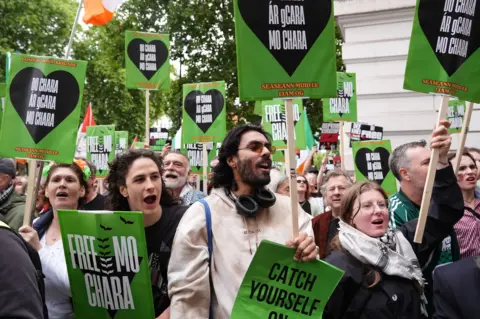PA Media A group of people gather holding green posters which say 'free mo chara' on them