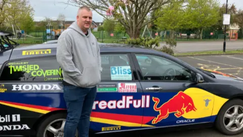 A man in a grey hoodie and blue jeans is stood alongside a grey estate car which is covered in different coloured logos and brand names