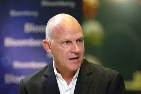 Campbell Wilson, wearing a black suit and a white shirt, speaks at a conference with a blue board behind him