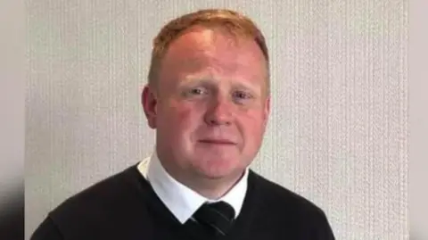 Legacy Independent Funeral Directors Robert Bush, undertaker, sits looking at the camera. He has short ginger hear and wears a black jumper