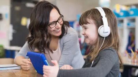 Teacher and child in a classroom. The child is wearing headphones and holding an ipad. They are both smiling. 