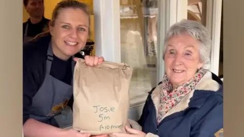 Josie collects her pasty from Gear Farm Pasty shop. She has grey hair and is wearing a scarf and wearing a navy jacket.
