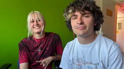 A young woman with blond hair and young man with brown, curly hair sit in a room with bright green walls. The young woman wears a burgundy top with a intricate black pattern inspired by heavy metal artwork and a silver pendant on a long chain.