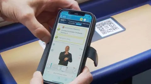 A person holds their iPhone while sitting on a train, it has the South Western Railway app open and the screen shows a video in the middle of the screen with a man who is about to use British Sign Language to explain an announcement