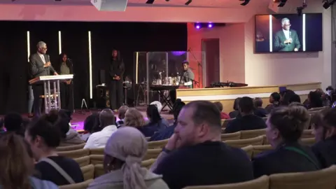The vigil service from the back of what looks similar to a lecture theatre. There is one speaker who is also projected on to a screen, and spectators on benches.