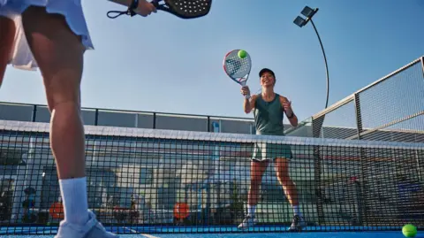 Two people playing padel on a court. A green ball is in mid-air as a woman who is smiling and wearing a green vest, shorts and trainers is holding up a racket and about to hit it. A net is between them with another woman standing in front of it wearing white trainers, socks and a skirt.
