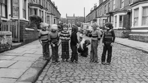 John Dean Black and white 1976 photograph of eight children standing across a cobbled street, with two pulling each other over