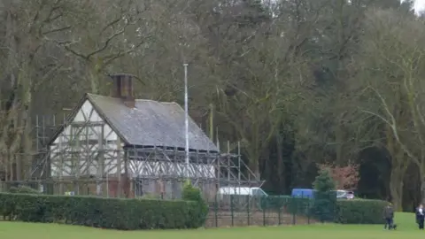 Elliott Brown A black and white timbered building covered in scaffolding and surrounded by grass, hedges and trees
