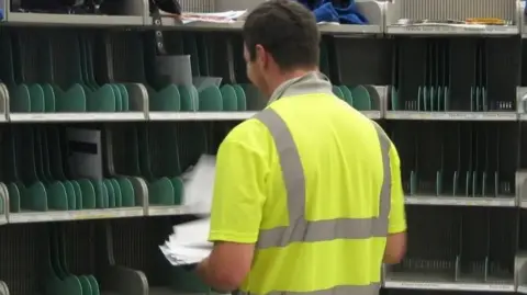 BBC A postal worker wearing a yellow hi-vis with his back facing the camera. He is sorting post onto the shelves which have green dividers. 