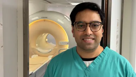 Dr Abhishek Sheth (dark hair, glasses, wearing green scrubs) looking at the camera. A large MRI scanner is behind him.