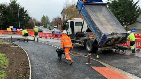 Workmen in orange and yellow high-visibility clothing working on the Longfield Gyratory in Trowbridge. A tipper truck with its tail raised is clearly visible, with a worker pushing a wheelbarrow away from the camera. A wall of plastic orange fencing is protecting the workforce.