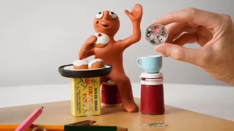 Alistair Heap/PA Wire A figurine of Aardman character Morph is sat down eating a belgian bun and waving his hand. He appears to be sat next to a small bottle of paint, colouring pencils and drawing pins. A human hand is holding up a 50p coin with Aardman characters on it including Wallace and Gromit and Shaun the Sheep.
