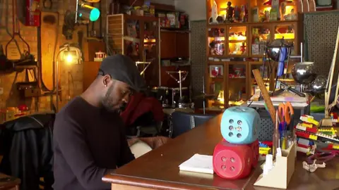 Owner Osho sits and plays the piano in the music school. He is wearing a flat cap and a long-sleeved black top. He is surrounded by trinkets and items collected over the years, including giant dice and a cabinet of various collectables.