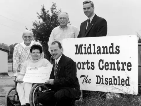 Getty Images A black and white image of a woman in a wheelchair holding a giant cheque. Len Tasker stands behind her. Three other men can be seen next to a giant sign that reads: Midlands Sports Centre for the Disabled.