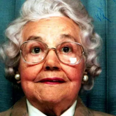 PA Media Mabel Leyshon, with a white curly perm and wearing glasses, in a passport style photograph. She wears pearl earrings and you can just see the collar of her white shirt in the photo with brown jacket lapels. 