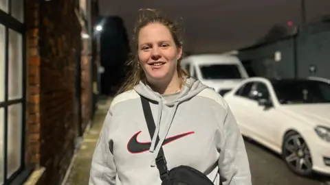 Anastasia smiles at the camera, her blonde hair is pulled back from her face, and she has a lip piercing. She wears a grey nike hoodie and a black crossbody bag.