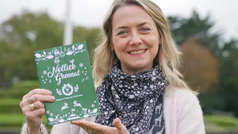 Culture Vannin A woman with blonde hair wears a black-and-white, star-decorated neck scarf and a white cardigan. She is holding a green book with the words Nollick Ghennal printed on it. She is outside.