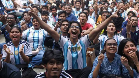 Getty Images Football fans watch the 2026 FIFA World Cup match between Argentina and Jordan on a giant screen in the Dhaka University area of Dhaka, Bangladesh, on 28 June. Many of them are wearing Argentina's iconic blue and white shirt