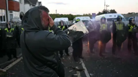 Getty Images A man in a hooded jacket is pictured clasping his hand to his face. Behind him is a row of police officers holding riot shields. 