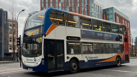 A red, blue and white double decker bus drives past a multi-storey building.