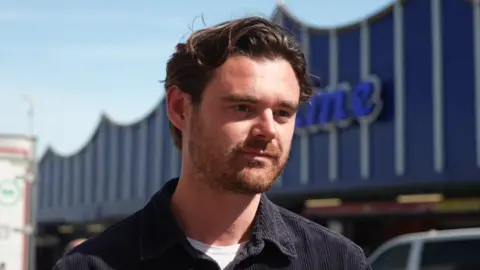 A man on a sunny day. He is standing outside an amusement arcade