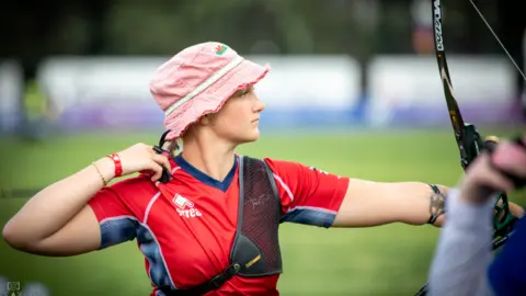 Turkish Archery Federation A woman with a red top and a black shoulder support with a pink beanie hat, holding a black bow with a green background