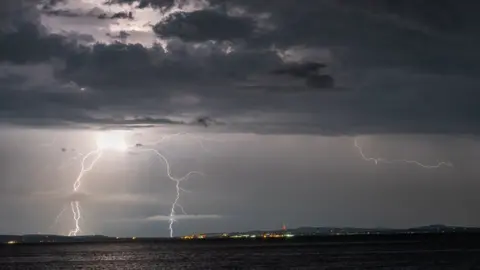 Shows grey skies with bolts of lightning above water and lights and mountains in the background