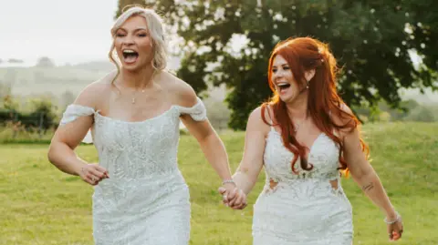 Lauren Price and Carlie Jones walking hand in hand in a field both laughing in white gowns