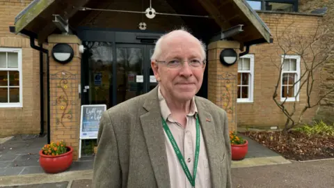 Paul Marriott standing in front of the entrance to St Cuthbert's Hospice. He is bald with short grey hair at the sides and is wearing glasses and a tweed blazer and khaki shirt. The building behind him is made from brick and has glass doors, tinted black.