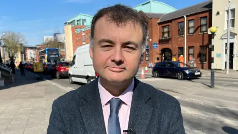 Gary Ridley looks at the camera. He is standing in front of a road and is wearing a blue suit jacket, blue tie and pink shirt.