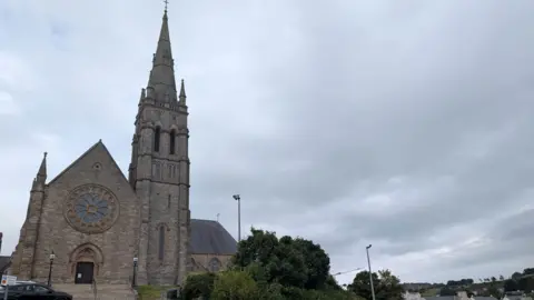 St Patrick’s Church in Downpatrick. There is a black car parked in front of it. The church is a large brown building, with a circular window in the front. The sky is overcast. 