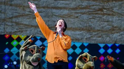 BBC / Sarah Jeynes / Jamie Simonds A wide shot of Matthew Murphy, the lead singer of The Wombats. He is wearing an orange shirt and singing into a microphone.