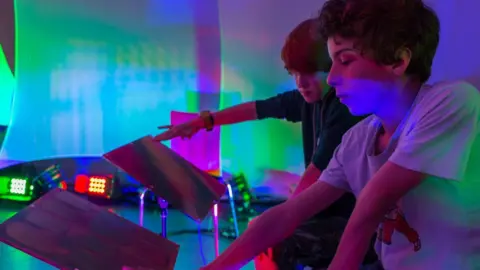 Two young boys in a room with multi coloured lights pressing buttons on a stand.