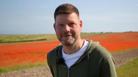 Shaun Whitmore/BBC Alan Percy, a man standing in front of a field of poppies at The Kelling Estate. He is looking directly at the camera and smiling. He is wearing a white T-shirt and green zip-up hoodie.