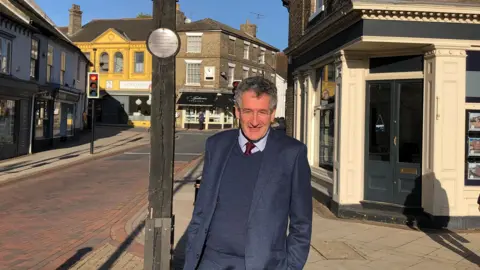 BBC/Vikki Irwin Suffolk MP Peter Prinsley wearing a tie and blue suit standing in the street. He has short dark curly hair and is next to a wooden post with a metal plaque on it. 