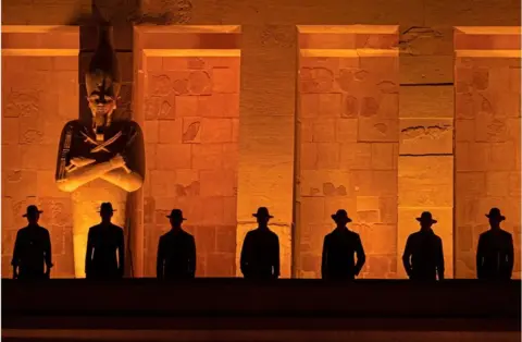 AFP Male models posing in front of an ancient Egyptian building. You can just see their black silhouettes.