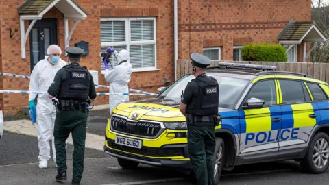 Police and forensic officers outside a red brick house. Police car is parked outside also. 