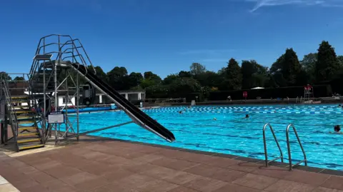 A bright blue outdoor swimming pool under a clear blue sky. There is a black slide into the pool and a ladder. There are people swimming in the pool, at the bottom end of which there is a white poolhouse. There are trees surrounding the site the lido is on.
