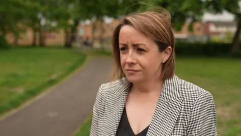 A woman with short brown hair in a chequered black and white jacket stands in a park 