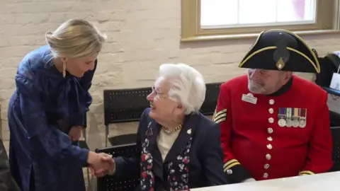 The Duchess of Edinburgh shakes hands with an older lady who is sitting at a table. A Chelsea Pensioner is sitting next to the lady, who has styled white hair. 