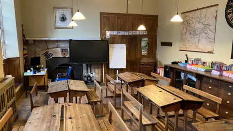 A photo which shows one of the temporary classrooms. There are four wooden desks in the photo with wooden benches tucked underneath them. In the middle of the photo there is a TV screen presumably used for teaching. Next to it there is also a whiteboard, and a map on the right-hand-side on the wall. 