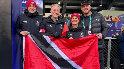 Axel Brown A group of four individuals standing together indoors at a winter sports venue. They are all wearing matching dark athletic jackets, some with red accents and national symbols visible on the sleeves and chest. A large red, black, and white flag of Trinidad and Tobago is being held across the front by members of the group.