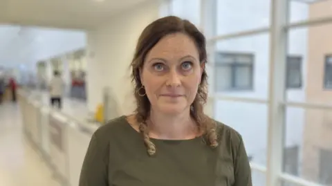 Claire Radley , Director of People, Gloucestershire Hospitals NHS FT. She is wearing a green top and is stood in a white corridor.